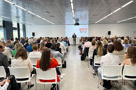 Das Bild zeigt einen großen Vortragsraum mit vielen vollbesetzten Stuhlreihen. Ganz vorne steht Prof. Weber mit einem Mikrofon und spricht zum Publikum.