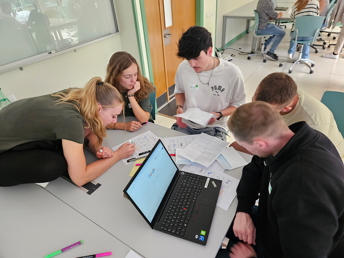 Fünf Studierende arbeiten gemeinsam an einem Tisch mit Unterlagen und Laptop in einem modernen Seminarraum.