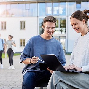 Foto einer Studentin und eines Studenten, die im Freien vor einem Gebäude sitzen und sich gemeinsam etwas auf einem Laptop ansehen. 