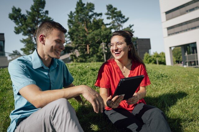 Foto einer Studentin und eines Studenten, die auf einer Wiese sitzen aund lachend auf ein Tablet gucken