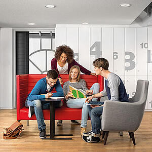 Zwei Studierende sitzen auf einem roten Sofa und lesen. Hinter dem Sofa steht eine Studentin. Rechts vom Sofa sitzt ein Student auf einem grauen Sessel und spricht.