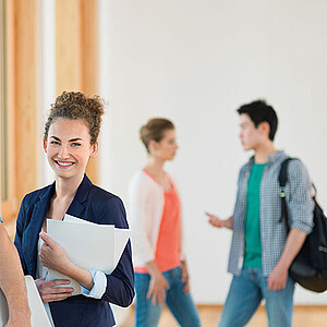 Eine Studentin steht im Vordergrund und blickt in die Kamera. Im Hintergrund unterhalten sich zwei Studierende.