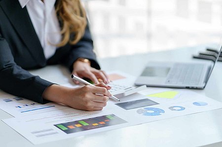 Schreibtisch mit Blättern an dem eine Person sitzt. Das Bild ist stark reingezoomt und die Person nicht ganz erkennbar. 