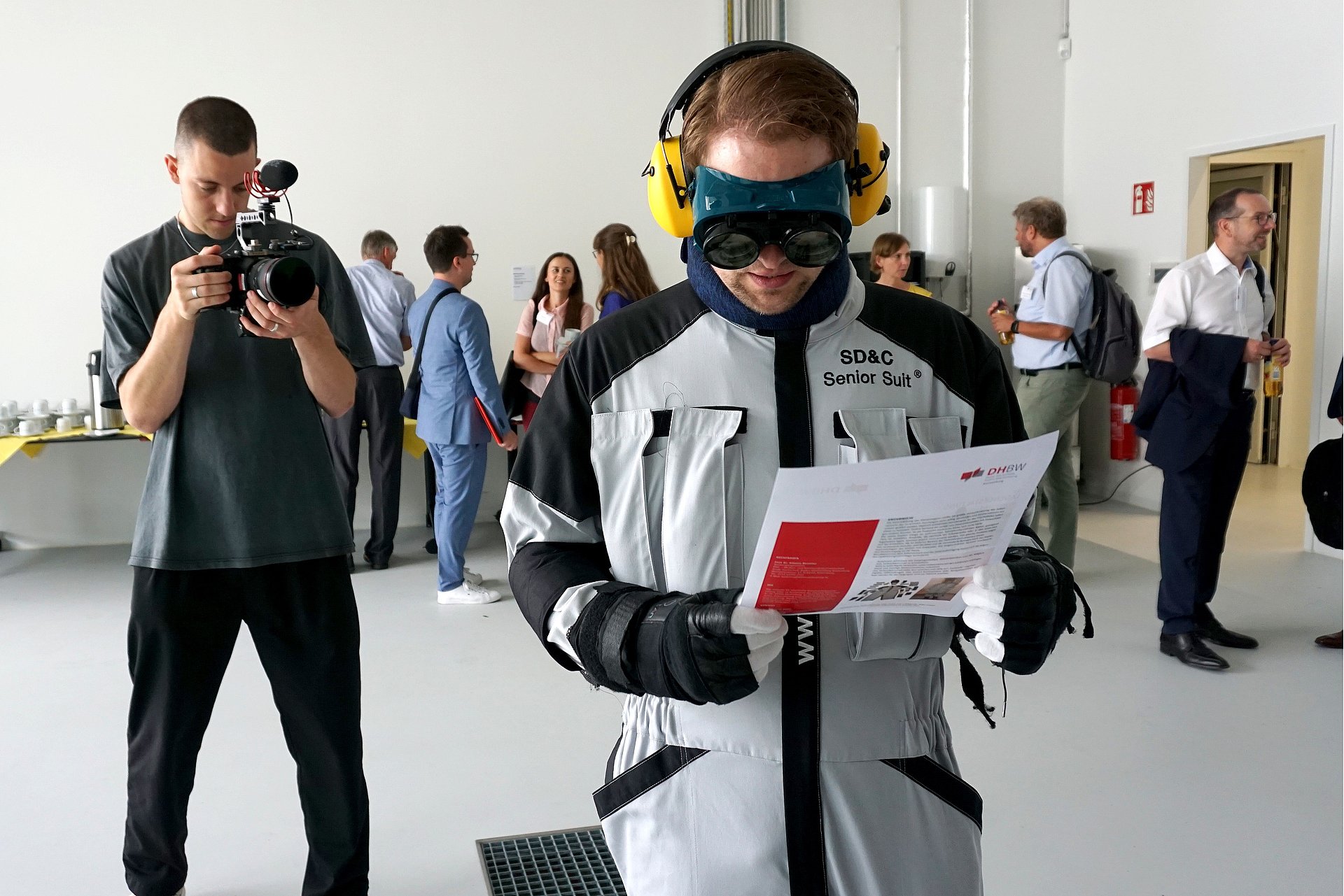 Foto einer Person, die einen Age Suit ausprobiert und versucht ein Dokument zu lesen. Im Hintergrund steht eine Person, die diese Szene fotografiert.