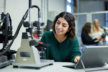 Junge Frau arbeitet konzentriert an einem Mikroskop in einem Labor und nutzt dabei ein Tablet zur Auswertung.