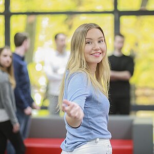 Foto einer blonden Studentin, die in die Kamera blickt und den Betrachtenden die Hand hinhält, als ob sie sie mitnehmen wollte