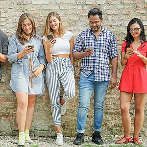 Foto von 4 jungen Personen in sommerlicher Kleidung, die vor einer Mauer stehen und auf ihre Smartphones schauen 