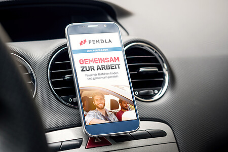 Smartphone in einer Halterung im Auto zeigt die Pendla-App mit dem Slogan ‚Gemeinsam zur Arbeit – passende Mitfahrer finden und gemeinsam pendeln‘.