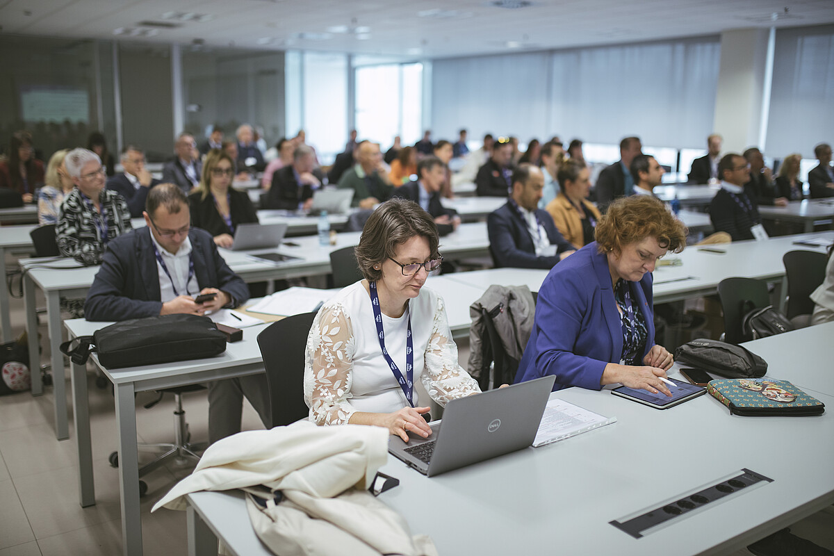 Blick in einen Seminarraum, an weißen Tischen Konferenzteilnehmer*innen, viele am Laptop.
