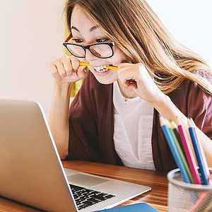 Foto einer jungen Frau mit Brille, die vor einem Laptop sitz und auf einen Stift beisst. 