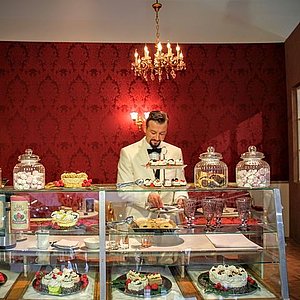 Foto eines Kellners in weißem Anzug, hinter einer Vitrine mit Torten, Kuchen und Keksen.