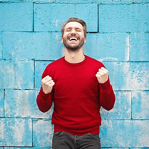 Foto eines Mannes in einem roten Pullover vor einer blauen Mauer, der die Hände zu Fäusten ballt und lacht.