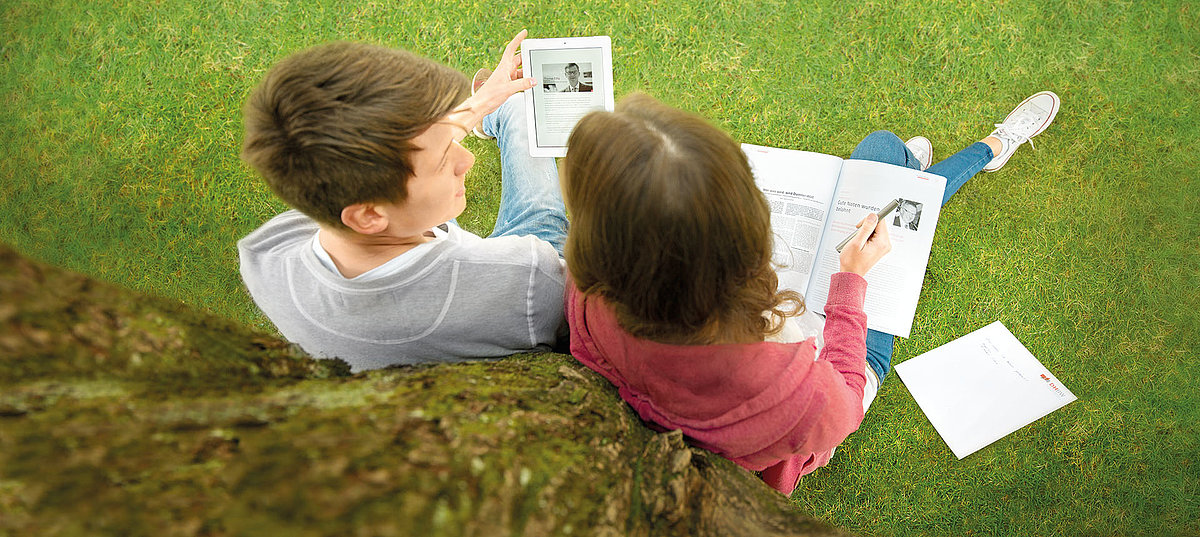 Ein Student (links) und eine Studentin (rechts) sitzen draußen auf dem Gras an einen Baum gelehnt. Man sieht sie von hinten / oben. Der Student hält ein Tablet in der Hand und die Studentin liest das Hochschulmagazin d13. 