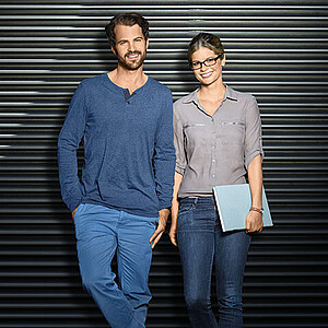 Ein Masterstudent (links) und eine Masterstudentin (rechts) schauen frontal in die Kamera. Er trägt einen Bart, sie trägt eine Brille und hält Unterlagen in der linken Hand. 