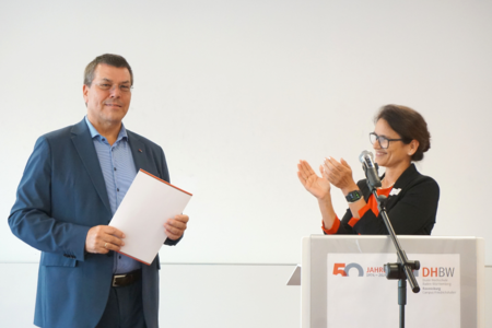 Herbert Dreher mit Urkunde in den Händen links neben Martina Klärle, die hinter einem Rednerpult steht und ihm applaudiert