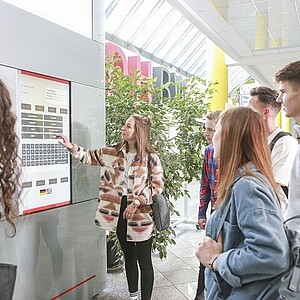 Foto einer Gruppe Studierender, die vor einem Info-Monitor stehen und Informationen zu Einrichtungen aufrufen.