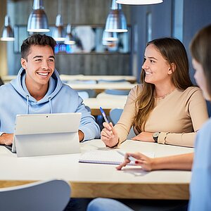 Foto von lachenden Studierenden, die gemeinsam an einem Tisch sitzen. Einer der Studierenden hat ein Tablet vor sich stehen.