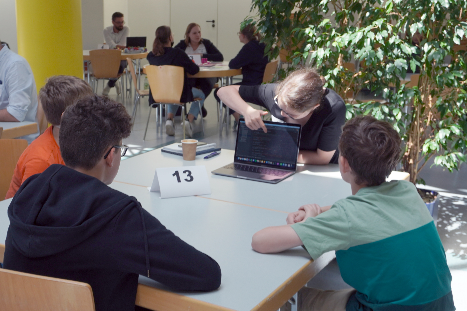 Drei Kinder sitzen an einem Tisch mit einer Mentorin die etwas erklärt