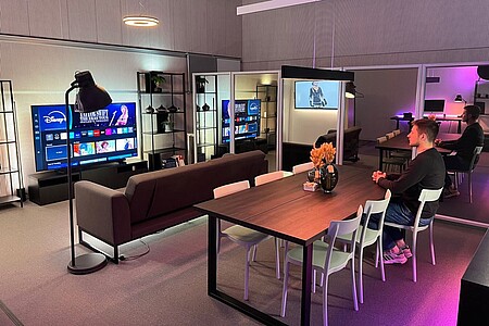 Blick in das Living Lab: rechts sitzt ein junger Mann an einem Tisch und schaut auf einen Fernseher, vor dem ein Sofa steht.