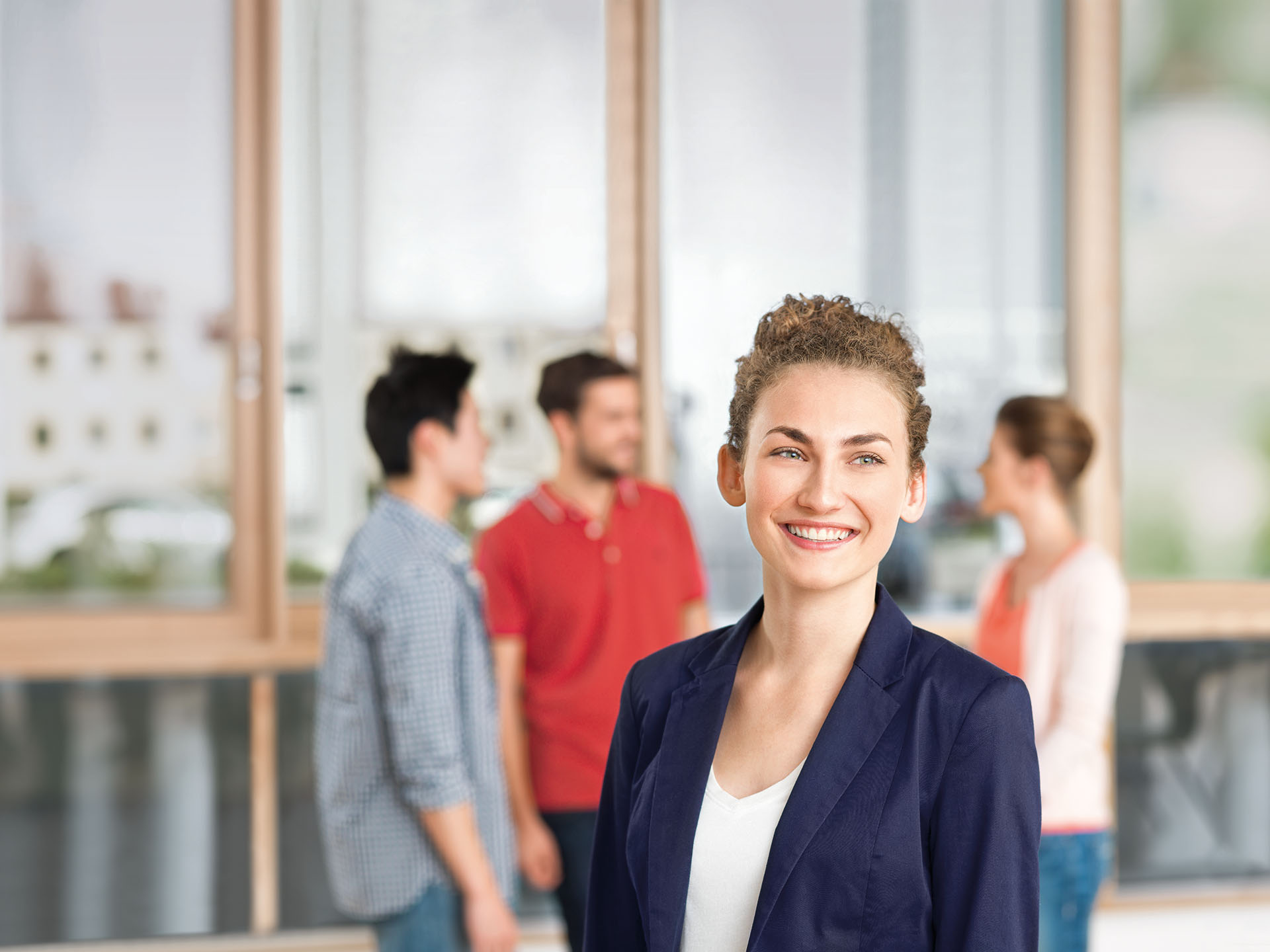 Eine Studentin steht im Vordergrund und blickt leicht nach rechts. Im Hintergrund unterhält sich eine Gruppe Studierender.