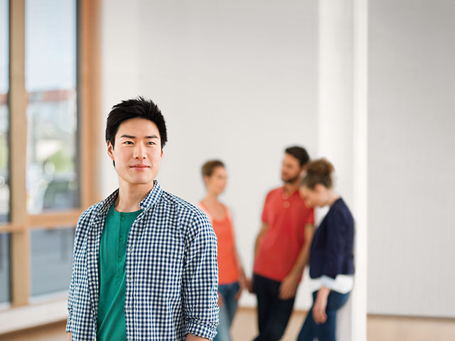 Frontalaufnahme eines asiatischen Studenten. Im Hintergrund unterhält sich eine Gruppe Studierender.