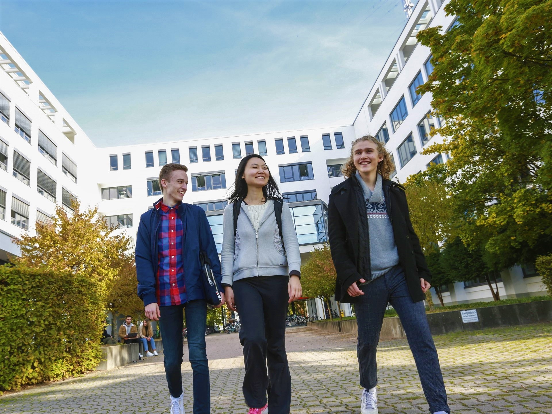 Foto von drei lachenden Studierenden mit einem weißen Gebäude und Bäumen im Hintergrund