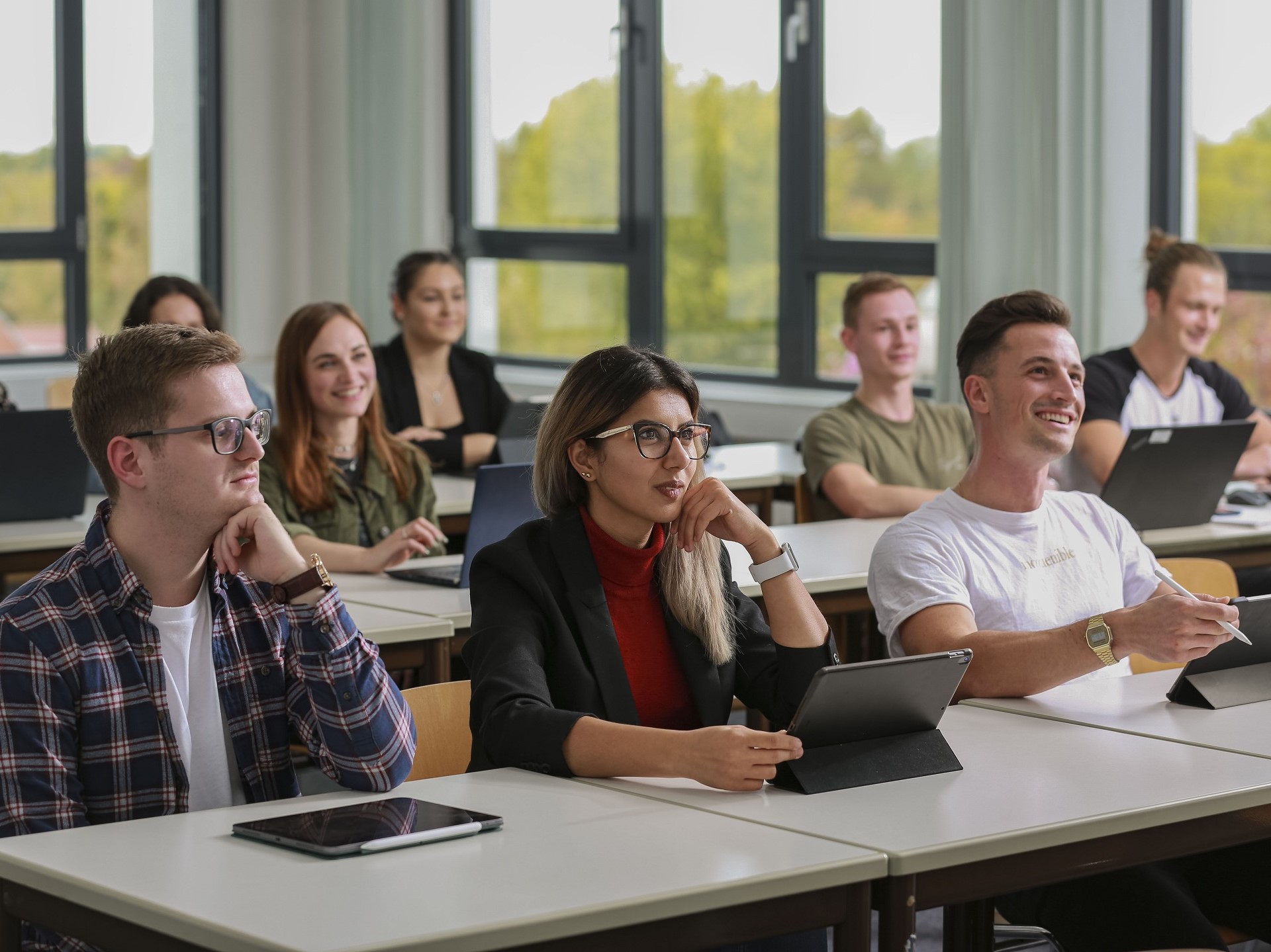 Foto von mehreren Studierenden in einem Vorlesungsraum. Einige haben Laptops oder Tabelts vor sich stehen. 