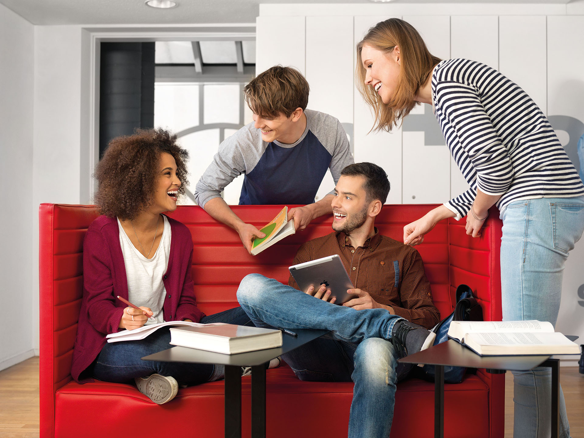 Ein Student und eine Studentin sitzen auf einem roten Sofa und unterhalten sich mit einem weiteren Studenten und einer Studentin.