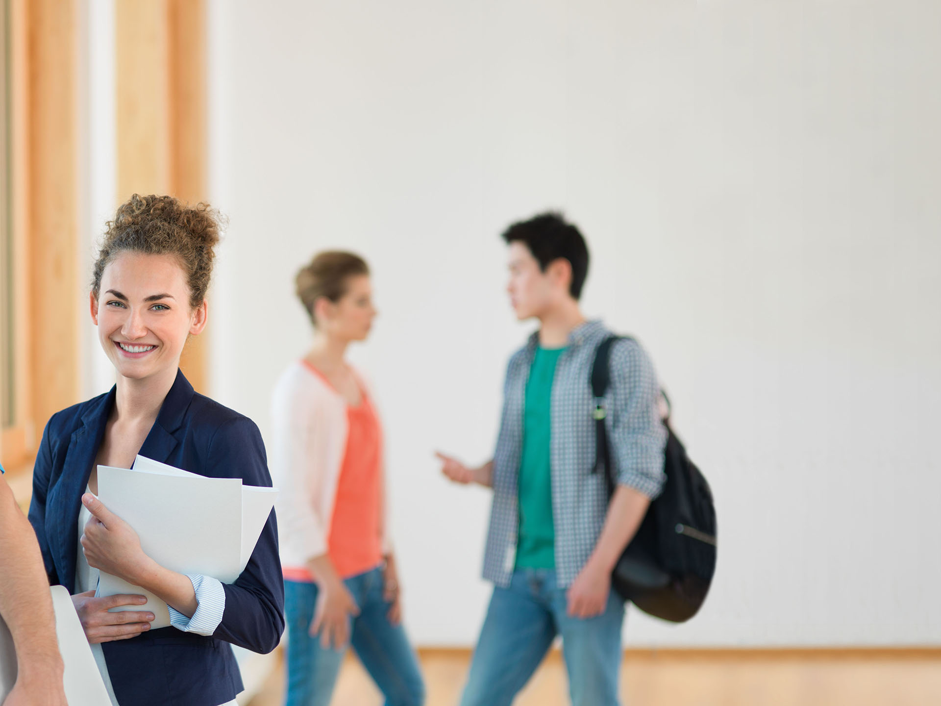 Eine Studentin steht im Vordergrund und blickt in die Kamera. Im Hintergrund unterhalten sich zwei Studierende.