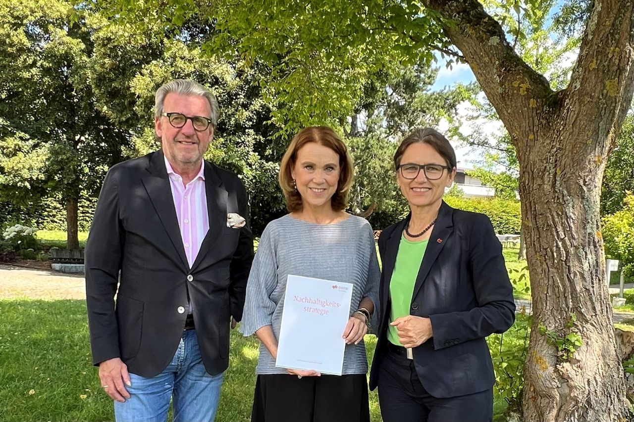 Von links nach rechts stehen im Grünen unter einem Baum Dr. Stefan Wolf, Petra Olschowski und Prof. Dr. Martina Klärle. Zweitere hält einen Ausdruck der Nachhaltigkeitsstrategie in den Händen.