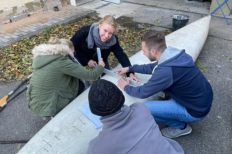 Zwei Frauen und zwei Männer basteln auf dem Boden vor einem Gebäude an einem Kanu aus Beton