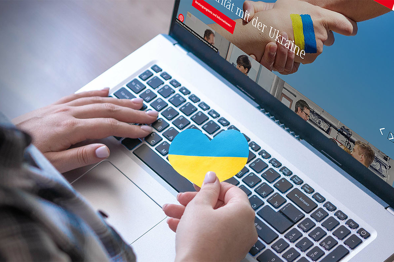 Blick von oben auf einen laptop mit der DHBW Website. Eine Hand hält ein Herz in Urkaine Farben vor die Tastatur.