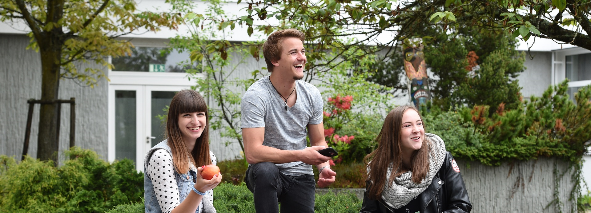 Foto von drei fröhlich lachenden jungen Personen vor einem Gebäude uns Pflanzen im Hintergrund. Eine Person lacht direkt in die Kamera.