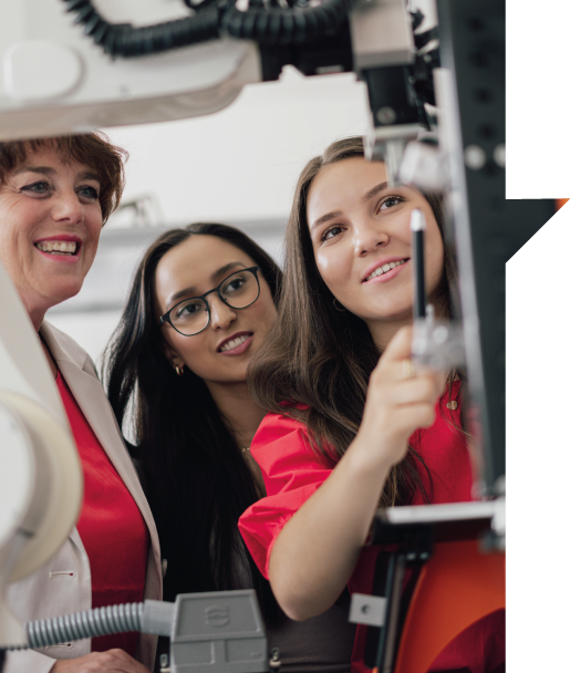 Zwei Studierende besprechen gemeinsam mit einer Professorin eine technische Maschine, eine der Studierenden zeigt mit einem Stift auf einen Bereich der Maschine.